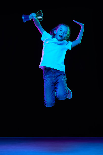 小美丽的女孩的肖像，孩子在白色t恤与奖杯和跳跃孤立的黑色工作室背景在霓虹灯。赢得图片下载