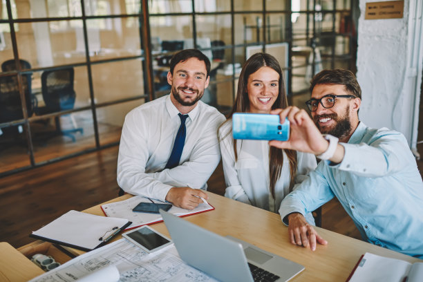 快乐的男女同事在室内用手机前置摄像头一起自拍，快乐的成功伙伴在办公室休息时用智能手机自拍图片下载