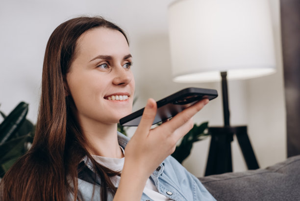 近距离的年轻美丽的女人使用语音短信或语音通信的手机，而坐在家里舒适的沙发上，微笑享受愉快的远程谈话的男朋友。技术概念图片下载