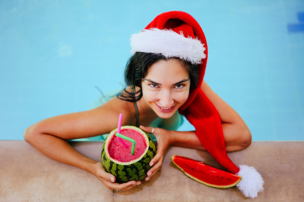 戴着圣诞帽的圣诞女人在游泳池里喝着西瓜鸡尾酒图片下载