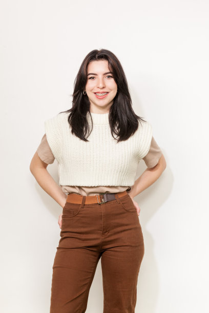 一个快乐的20岁白人女性的肖像，黑色长发，白色针织马甲和白色背景的棕色裤子图片下载