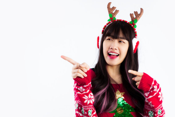 身穿红白绿相间运动衫的亚洲妇女戴着耳罩和驯鹿角头巾，在白色背景上摆出手指指向的姿势。圣诞快乐。图片下载