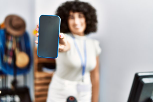 年轻的中东女店主微笑着开心地在服装店展示应用程序智能手机。图片下载