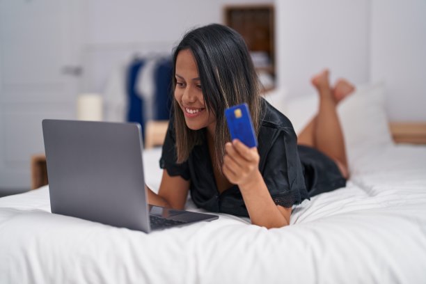 年轻的西班牙妇女用笔记本电脑和信用卡躺在卧室的床上图片下载