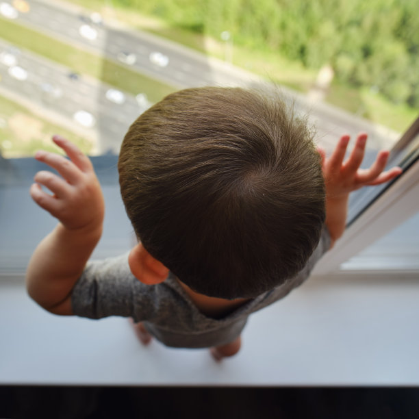 蹒跚学步的婴儿正站在窗边的窗台上。窗户边的孩子双手扶着玻璃。一岁儿童图片下载