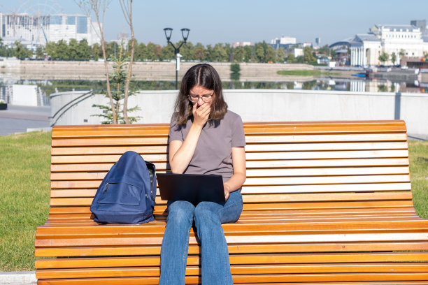 疲惫的学生女孩坐在公园的长凳上，在笔记本电脑上打字。这女孩工作过度睡着了。图片下载