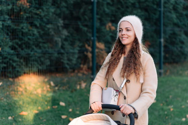 身穿米色外套的年轻女子推着婴儿车里的孩子穿过小镇。妈妈带着婴儿车。一位推着婴儿车的妇女在日落时分走在公园里。图片下载