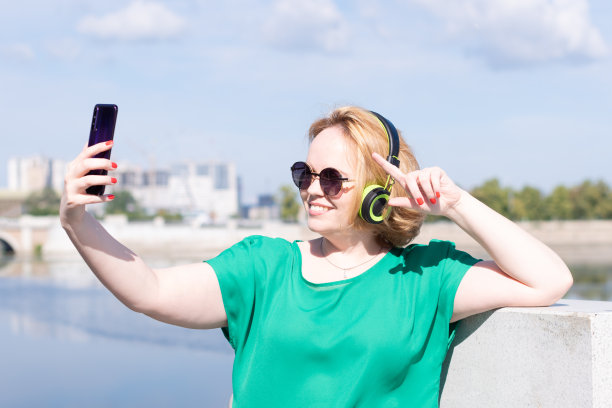 一个快乐的女学生戴着墨镜和耳机在手机上自拍，在街上和朋友聊天。图片下载
