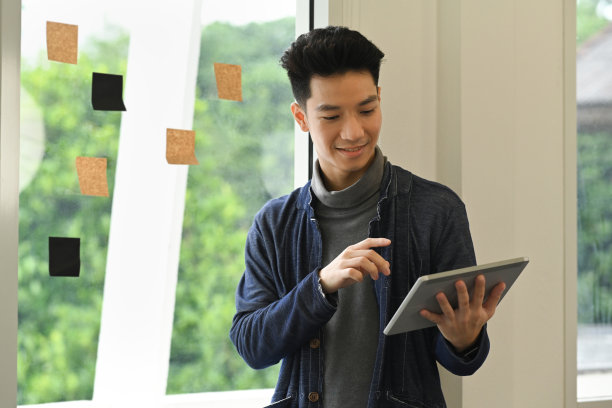 微笑着年轻有创意的男性自由职业者使用数字平板电脑，而站在窗口附近与便签。图片下载