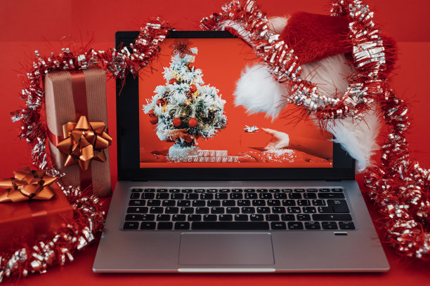 桌子与笔记本电脑在圣诞节时间，事件，节日，庆祝，圣诞快乐的消息红色背景与圣诞礼物和圣诞老人帽子图片下载