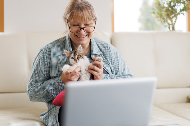 穿着蓝色衬衫的Marure女士坐在家里的木桌上，腿上坐着一只猫，手里拿着笔记本电脑和笔记本电脑，在网上工作或购物。图片下载