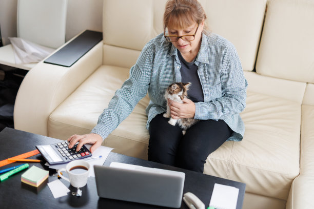 穿着蓝色衬衫的Marure女士坐在家里的木桌上，腿上坐着一只猫，手里拿着笔记本电脑和笔记本电脑，在网上工作或购物。图片下载