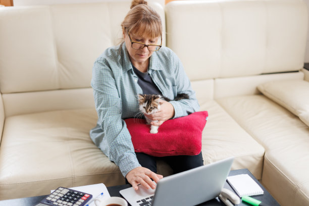 穿着蓝色衬衫的Marure女士坐在家里的木桌上，腿上坐着一只猫，手里拿着笔记本电脑和笔记本电脑，在网上工作或购物。图片下载