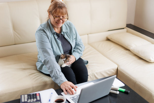 穿着蓝色衬衫的Marure女士坐在家里的木桌上，腿上坐着一只猫，手里拿着笔记本电脑和笔记本电脑，在网上工作或购物。图片下载