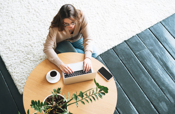 年轻微笑的女人在米色开衫和牛仔裤在房间使用笔记本电脑，俯视图图片下载