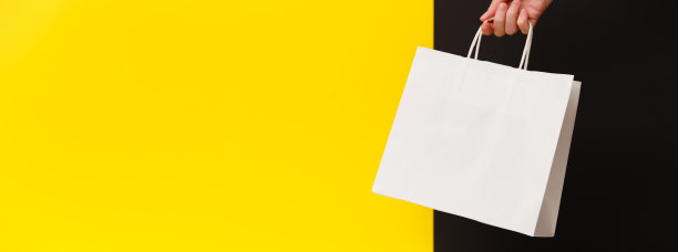 女性手拿白色空白购物袋在黄色和黑色的背景。白色工艺空白纸袋在手。折扣、购物和生态的概念。黑色星期五销售横幅。图片下载