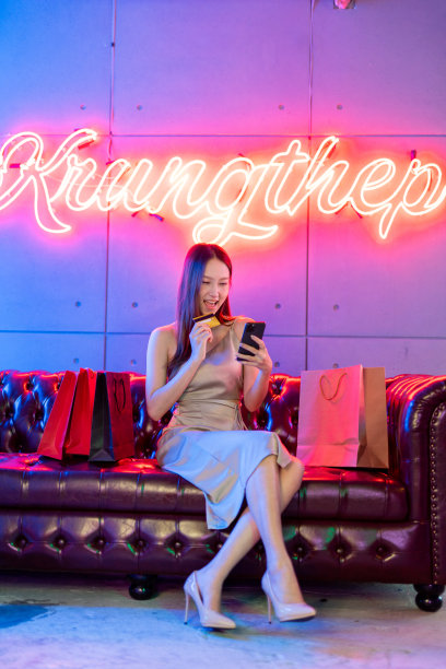 肖像照片的年轻美丽的亚洲女模特穿着优雅的衣服在霓虹灯的房间支付与她的信用卡借记卡在她的手机网上购物狂欢图片下载