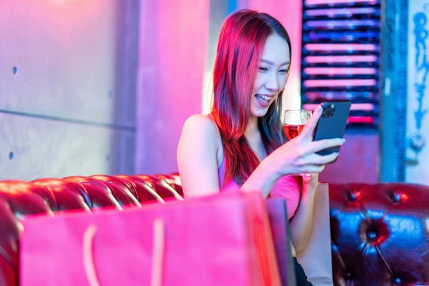 肖像照片的年轻美丽的亚洲女模特在霓虹灯房间聊天愉快的她的手机，同时有一杯酒后的购物狂欢图片下载