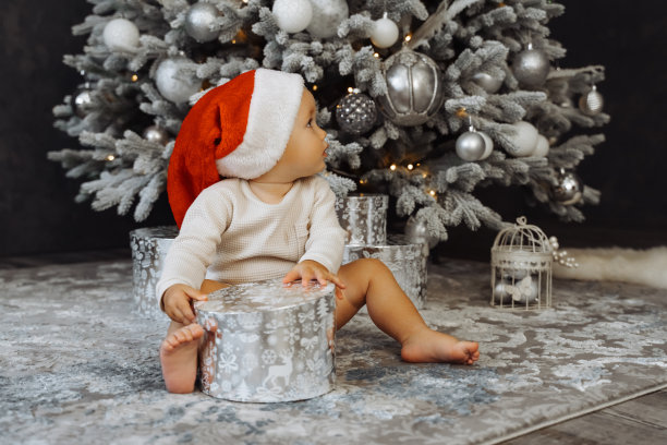 1岁的宝宝坐在圣诞帽上，房间里有圣诞树和背景灯。圣诞快乐，新年快乐。假期。图片下载