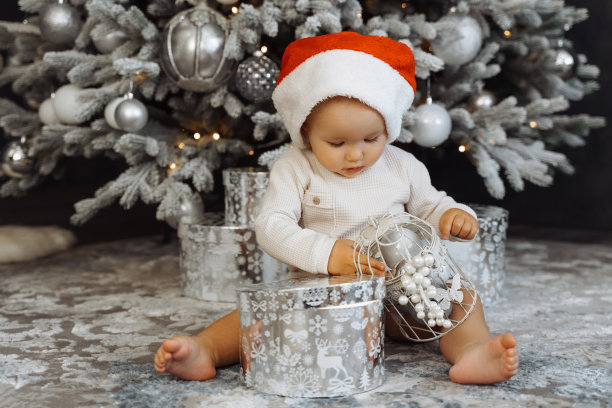 1岁的宝宝坐在圣诞帽上，房间里有圣诞树和背景灯。圣诞快乐，新年快乐。假期。图片下载