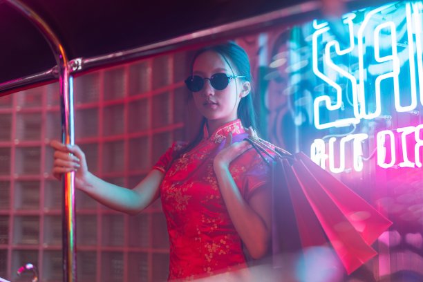 透过玻璃拍摄一位年轻的亚洲女模特，她戴着墨镜，穿着红色中国传统旗袍，在霓虹灯前看起来很酷，拿着购物袋，站在一辆嘟嘟出租车旁边图片下载