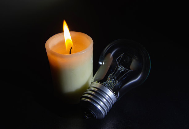 在完全黑暗中燃烧的蜡烛附近没有发光的灯泡。停电城市，停电，减载，能源危机或停电，象征性形象。图片下载