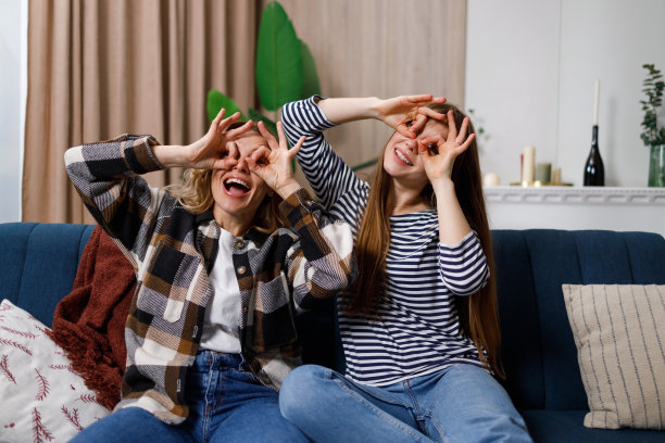 成熟的女人和成年的女儿嬉闹着做鬼脸用手指做望远镜，坐在家里的沙发上图片下载