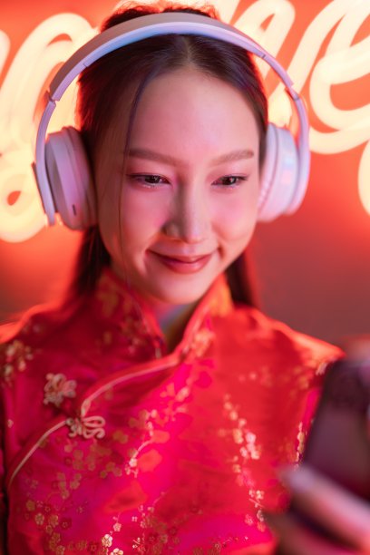 肖像照片的美丽年轻的亚洲女性模特可爱的样子，戴着耳机和红色传统中国旗袍，在橙粉色霓虹灯前听音乐使用她的手机智能手机图片下载