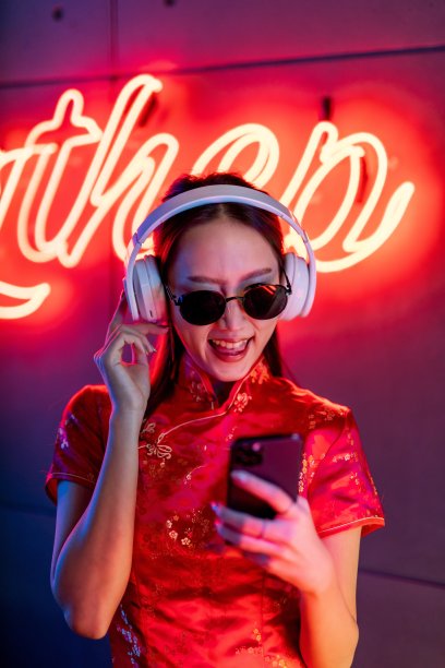 肖像照片的美丽年轻的亚洲女性模特可爱的样子，戴着太阳镜，耳机和红色传统中国旗袍，在橙粉色霓虹灯前听音乐使用她的手机智能手机图片下载