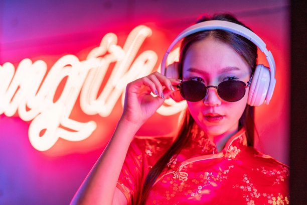 近距离的肖像照片，一个美丽的年轻的亚洲女性模特酷的样子，戴着太阳镜，耳机和红色的传统中国旗袍，在橙粉色霓虹灯前听音乐图片下载