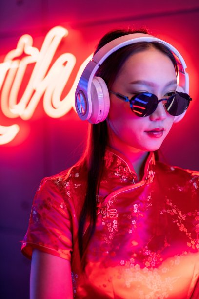 近距离的肖像照片，一个美丽的年轻的亚洲女性模特酷的样子，戴着太阳镜，耳机和红色的传统中国旗袍，在橙粉色霓虹灯前听音乐图片下载