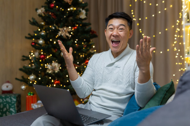 一个年轻的亚洲人坐在家里，拿着一台笔记本电脑，为圣诞节抽奖的胜利而高兴图片下载