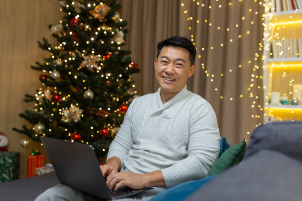 一个年轻的亚裔自由商人坐在家里的圣诞树旁，手里拿着一台笔记本电脑图片下载