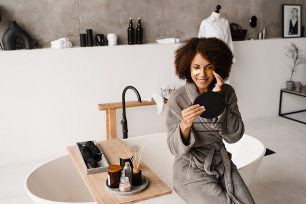 一位眼睛上有金色眼罩的非裔美国妇女看着镜子里她的皮肤。早晨美容例行程序。穿着浴袍的非洲女孩在浴室里把金贴片涂在眼睛上。图片下载