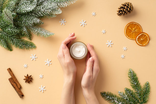 冬季护肤理念。第一人称俯视图照片的妇女的手奶油罐冷杉枝松果干橙片肉桂棒八角和雪花在孤立的柔和米色背景图片下载
