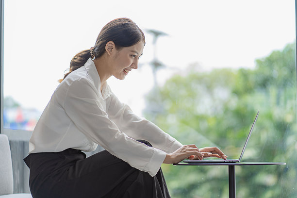 迷人的亚洲女子独自坐在咖啡馆的桌子旁，她看起来像是在看着自己的笔记本电脑工作，微笑着，工作在线的概念。图片下载