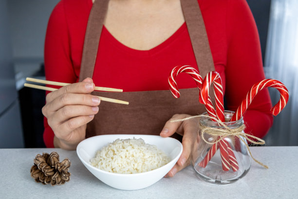 在昏暗的厨房里，一个穿着红衬衫和棕色围裙的女孩手里拿着一盘金色的米饭。桌上有一罐新年糖果和一个甜筒。亚洲菜和圣诞概念图片下载