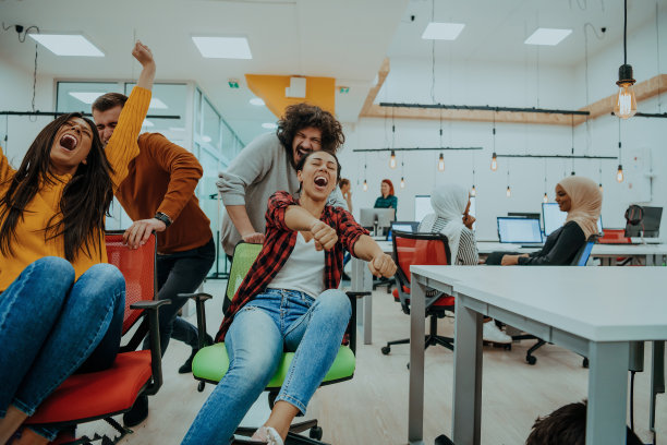 团队建设和办公室乐趣。年轻愉快的商务人士穿着时髦的休闲装，在办公椅上奔跑，微笑着。图片下载