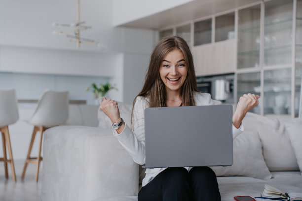 成功的年轻女企业家通过互联网与伙伴交谈，握紧拳头，像赢家，微笑着在家里做视频电话，坐在沙发上，使用笔记本电脑。商业和远程教育。图片下载