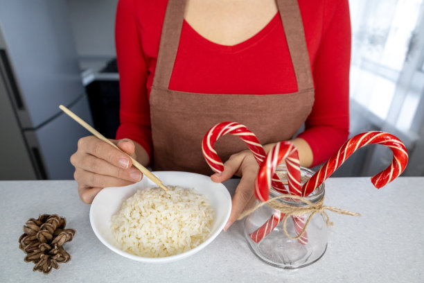 在昏暗的厨房里，一个穿着红衬衫和棕色围裙的女孩手里拿着一盘金色的米饭。桌上有一罐新年糖果和一个甜筒。亚洲菜和圣诞概念图片下载
