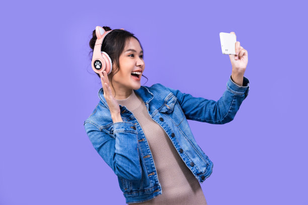 美丽的亚洲年轻女子在夹克牛仔裤和玩电子游戏使用操纵杆与紫罗兰色背景隔离的耳机。图片下载