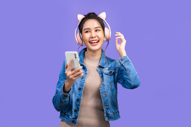 美丽的亚洲年轻女子在夹克牛仔裤和玩电子游戏使用操纵杆与紫罗兰色背景隔离的耳机。图片下载