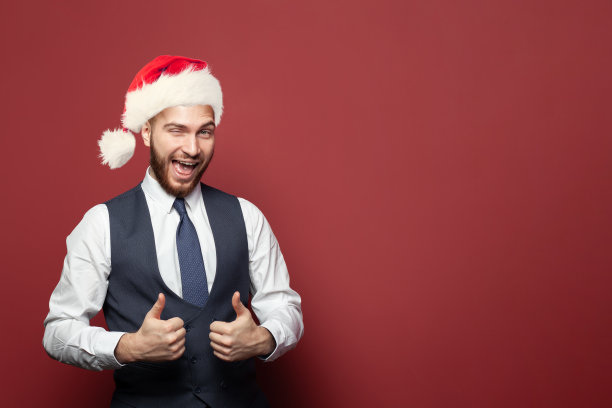 快乐的成功商人西装革面，戴着圣诞帽，在工作室红色的墙壁背景上眨眼，竖起大拇指图片下载