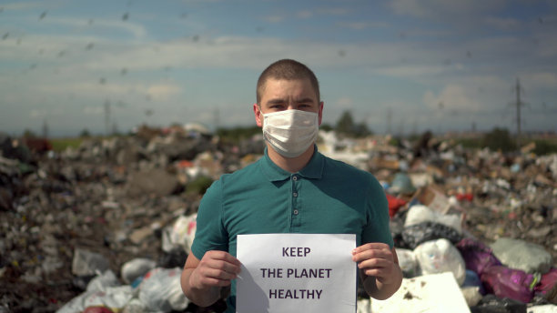 一名戴着医用口罩的年轻男子举着“保持地球健康”的海报站在垃圾场前。鸟儿飞向垃圾堆。图片下载