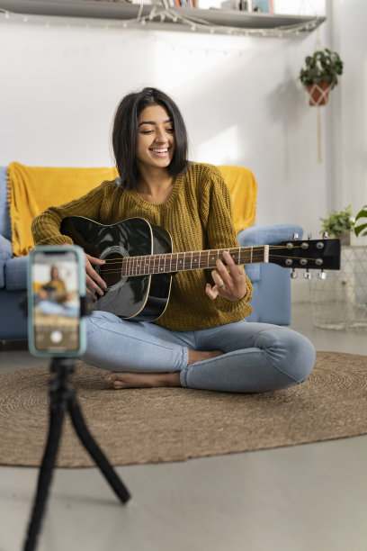 年轻的棕发印度女子一边用智能手机录制视频，一边弹着原声吉他。千禧年印度女性在家。图片下载