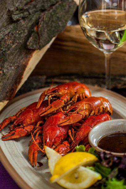小龙虾与香草、柠檬角和混合沙拉一起在沸水中煮熟，放在一个米色的椭圆形盘子里。旁边是一个米色的陶瓷碗，盛着洋葱酱。盘子放在紫色的桌布上，旁边是一杯白葡萄酒和几块木块。图片下载