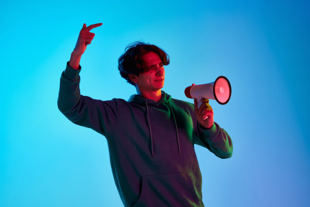 年轻男子卷发摆姿势与扩音器在霓虹灯蓝色背景孤立的肖像。学生会议图片下载