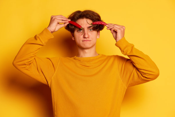 卷头发的年轻男子的肖像与辣椒孤立在黄色背景。扮鬼脸图片下载