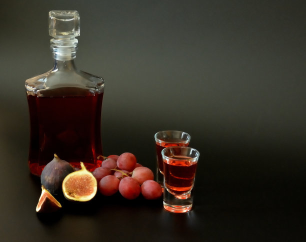 黑色背景的水果利口酒，一瓶和两杯自制酒精，旁边是几片成熟的无花果和一串深色葡萄。图片下载