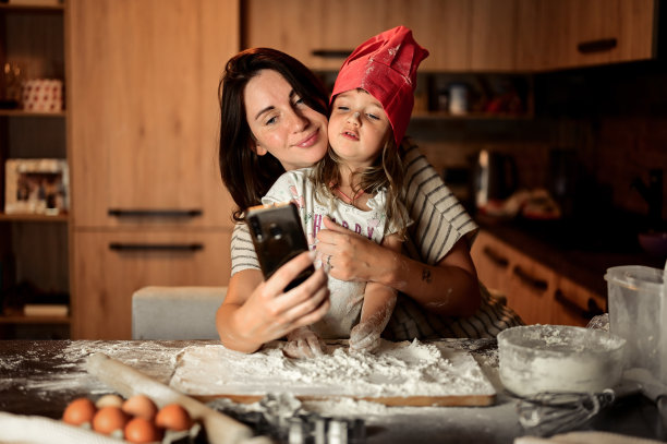 幸福家庭的母女在厨房里揉面。妈妈和小女儿在厨房一起烘焙时用智能手机自拍，图片下载